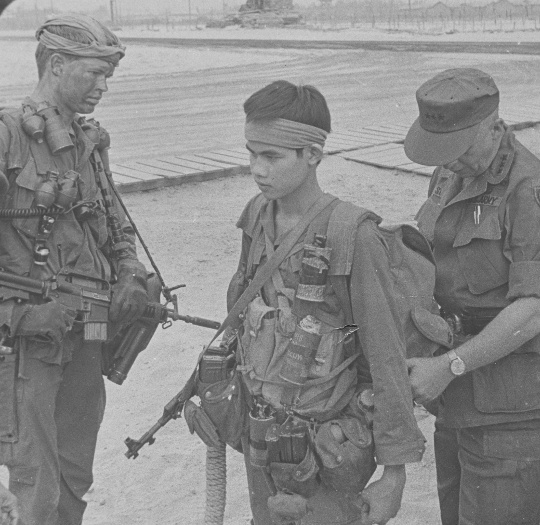 This April 1969 photo of an RT Idaho inspection at top secret CCN compound/headquarters in Da Nang by Lt. Gen. Richard Stilwell, the I Corps commander at the time, reflects the general attire worn by SOG recon men. On the left, is author John Stryker Meyer, holding SOG's primary weapon of choice, the CAR-15. Immediately behind it is the sawed-off M-79, 40 mm grenade launcher. On the right harness strap there are grenades and the conventional smoke grenade at the top with the smaller, white smoke grenades contained in aluminum containers. On his right hip is the survival axe. Son is the team member being inspected by Stilwell. Son was the pointman for RT Idaho at that time and is wearing an NVA uniform, the traditional NVA magazine vest across his stomach area, while carrying the AK-47, the NVA's weapon of choice. The rope dangling from his right side is used for Swiss seats, which is wrapped around the waist, between the legs and worn for extraction by ropes, dropped from hovering helicopters in areas where there isn't enough room for helicopters to land. Son has the 30-round NVA AK-47 magazines held in canvas canteen pouches and another canteen pouch holding frag grenades.